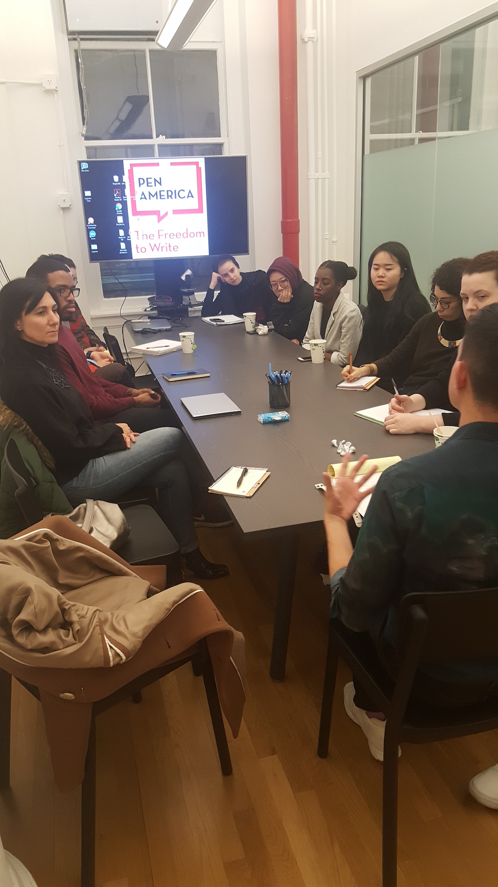 Students sitting along rectangular table with screen at the end reading Pen America